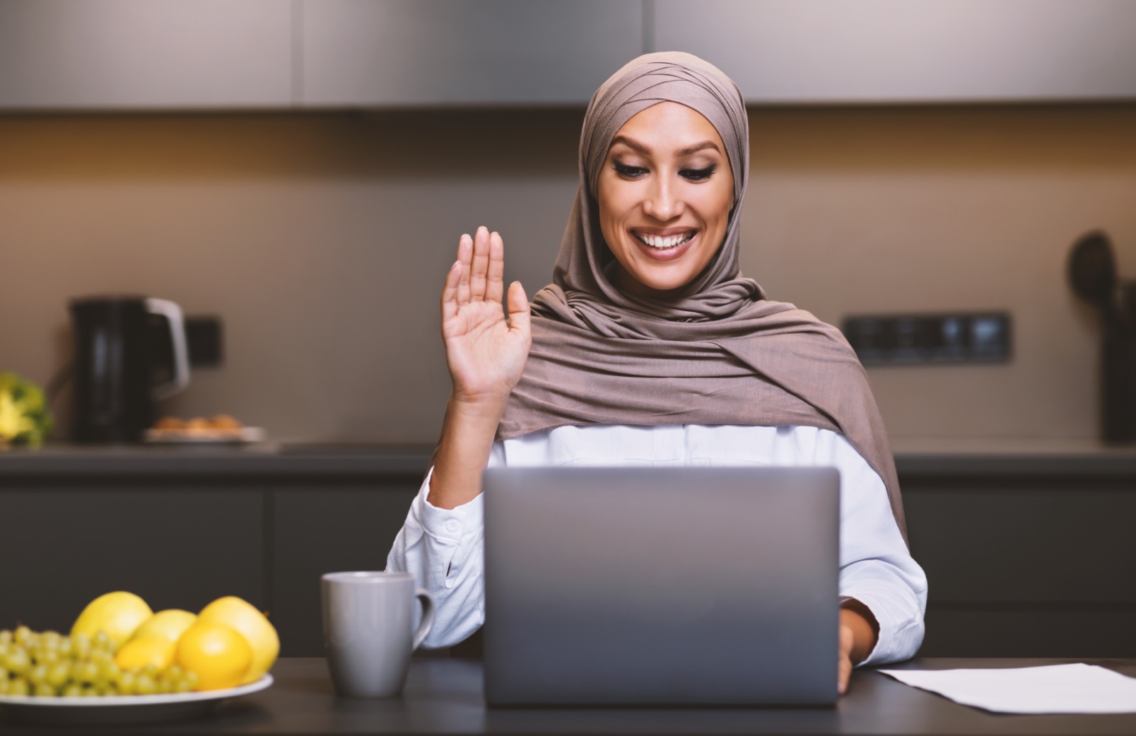 Muslim Lady At Laptop Making Video Call Waving Hello Sitting In Modern Kitchen At Home, Wearing Hijab. Arab Woman Using Computer Communicating Remotely. Female Dietitian Having Online Consultation.