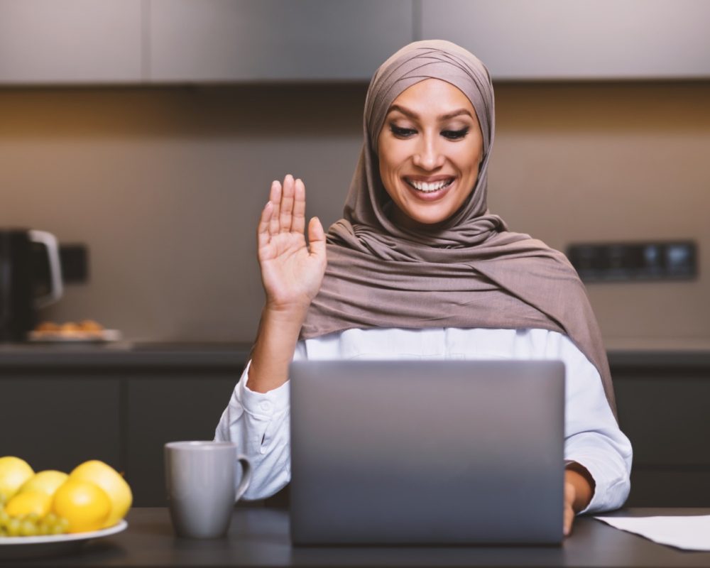 Muslim Lady At Laptop Making Video Call Waving Hello Sitting In Modern Kitchen At Home, Wearing Hijab. Arab Woman Using Computer Communicating Remotely. Female Dietitian Having Online Consultation.