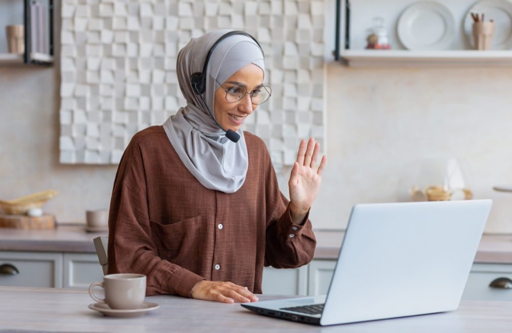 Woman in hijab working remotely from home sitting in kitchen using laptop and headset for video call, businesswoman smiling and talking to customers remotely customer support service online tech